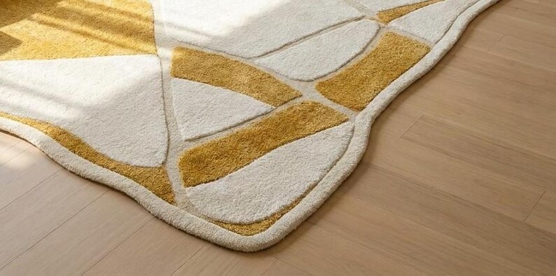 Pu&ograve; includere: Primo piano di un moderno tappeto con un design geometrico. Il tappeto presenta uno sfondo color crema con sezioni giallo senape. I bordi sono arrotondati e il tappeto &egrave; posato su un pavimento in legno chiaro.