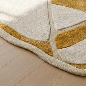 Pu&ograve; includere: Un tappeto color crema con un design geometrico con forme giallo dorato. Il tappeto ha un aspetto morbido e strutturato e un bordo curvo. Il tappeto &egrave; posto su un pavimento in legno chiaro.