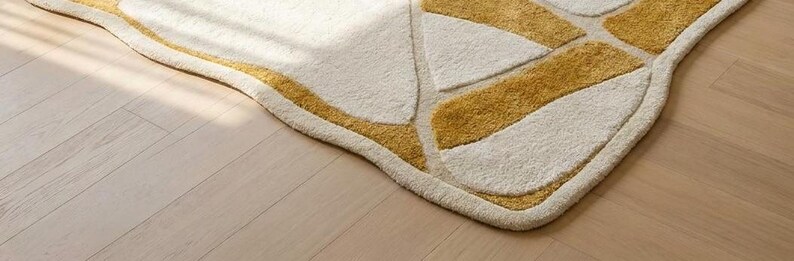 Pu&ograve; includere: Un tappeto color crema con un design geometrico con forme giallo dorato. Il tappeto ha un aspetto morbido e strutturato e un bordo curvo. Il tappeto &egrave; posto su un pavimento in legno chiaro.