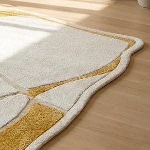 Pu&ograve; includere: Tappeto rettangolare color crema con un bordo curvo unico. Il tappeto presenta un design geometrico nelle tonalit&agrave; dell'oro e del crema, creando una superficie strutturata e invitante. Il tappeto &egrave; posato su un pavimento in legno chiaro.