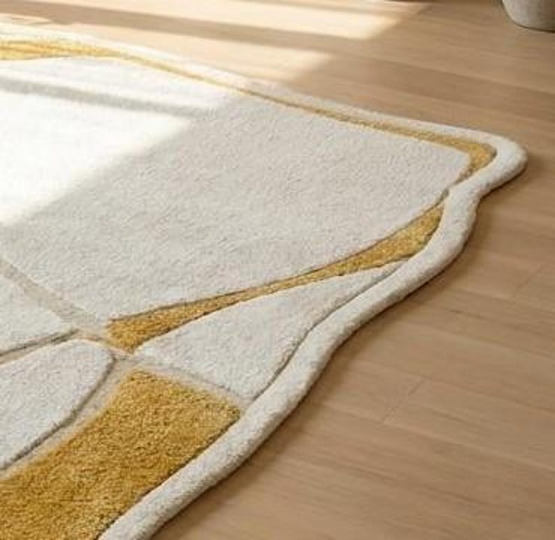 Pu&ograve; includere: Tappeto rettangolare color crema con un bordo curvo unico. Il tappeto presenta un design geometrico nelle tonalit&agrave; dell'oro e del crema, creando una superficie strutturata e invitante. Il tappeto &egrave; posato su un pavimento in legno chiaro.
