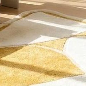 Pu&ograve; includere: Un tappeto bianco e oro con un design astratto. Il tappeto presenta una superficie morbida e strutturata con forme geometriche in oro e bianco. Il tappeto &egrave; posato su un pavimento in legno, con uno scorcio di un divano beige sullo sfondo.