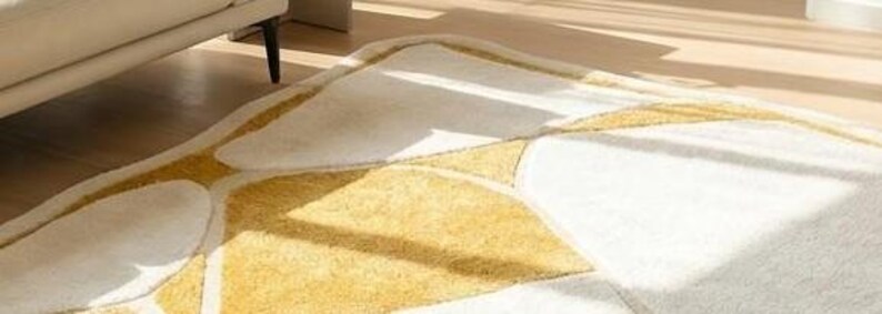 Pu&ograve; includere: Un tappeto bianco e oro con un design astratto. Il tappeto presenta una superficie morbida e strutturata con forme geometriche in oro e bianco. Il tappeto &egrave; posato su un pavimento in legno, con uno scorcio di un divano beige sullo sfondo.