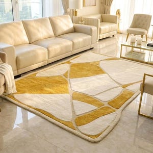 Pu&ograve; includere: Un soggiorno moderno con mobili color crema, tra cui un ampio divano, poltrone e un tappeto fantasia con forme geometriche gialle e bianche. Un tavolino in vetro con cornici dorate e dettagli dorati completano l'elegante design.