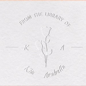 Könnte beinhalten: Ein weißer Prägestempel mit dem Text "From the Library of" in einem Kreis um eine stilisierte Rose. Der Text "Kim Arabella" befindet sich unter der Rose.