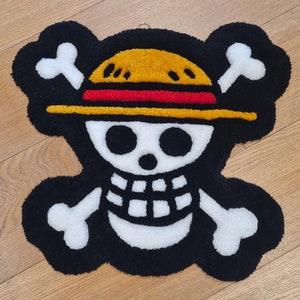 Puede incluir: Una alfombra negra y blanca con un diseño de calavera y huesos cruzados. La calavera lleva un sombrero de paja amarillo y rojo.