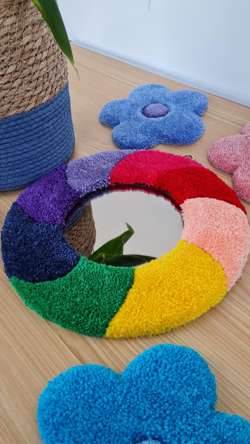May include: A round mirror with a rainbow-colored frame made of fluffy yarn. The mirror is surrounded by a variety of colorful, fluffy flower shapes.
