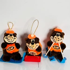 Könnte beinhalten: Drei Vintage-A&W-Root-Beer-Bären-Ornamente. Jeder braune Bär trägt ein orangefarbenes Hemd und einen Hut mit dem A&W-Logo. Ein Bär sitzt auf einem blauen Schlitten, einer auf einem roten Schlitten und einer auf blauen Skiern. Goldene Schlaufen zum Aufhängen.