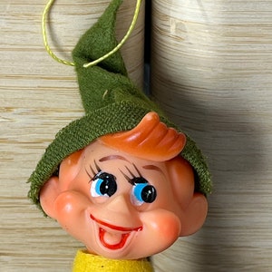 Puede incluir: Un gorro de elfo de fieltro verde con un cuello de fieltro amarillo y una cara sonriente con ojos azules. El elfo está hecho de plástico y tiene una boca roja.