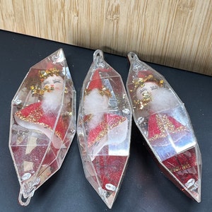 Op de afbeelding: Drie vintage kerstversieringen in de vorm van heldere, gefacetteerde diamanten. Elke versiering bevat een kleine, rood en goud geklede pop met een wit gezicht en gesloten ogen.