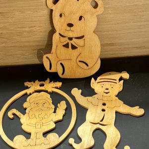 Op de afbeelding: Drie houten kerstversieringen: een beer, een kerstman en een elf. De versieringen zijn uit hout gesneden en hebben een natuurlijke houten afwerking.