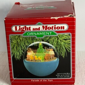 May include: A red box with a blue and white Christmas ornament inside. The ornament features a scene of children playing with toys around a Christmas tree. The box reads "Light and Motion Ornament" and "Parade of the Toys". The text on the box also says "Jack-in-the-Box bobs up & down as toys parade."