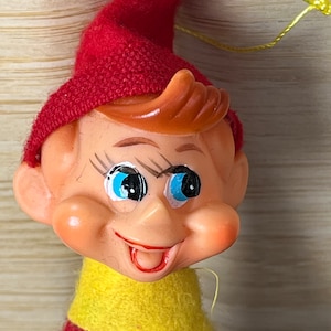 Puede incluir: Un adorno de elfo de fieltro rojo y amarillo con una cara sonriente y ojos azules. El elfo lleva un gorro rojo y una bufanda amarilla.