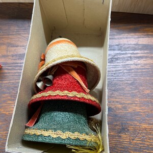 Puede incluir: Tres campanas de Navidad vintage en una caja de cartón. Las campanas están apiladas una encima de la otra, con la campana más grande en la parte inferior y la más pequeña en la parte superior. Las campanas están decoradas con purpurina, ribetes dorados y cintas de terciopelo rojo y verde.