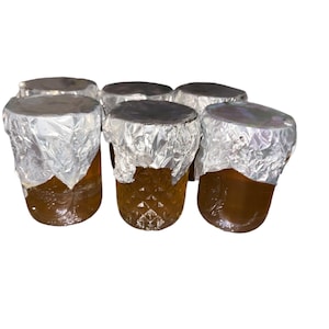Puede incluir: Seis pequeños frascos de vidrio con tapas de papel de aluminio plateado llenos de un líquido dorado. Los frascos están dispuestos en semicírculo.