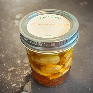 May include: A clear glass jar filled with fermented garlic honey. The jar has a silver lid and a label that reads "Green Grocer" and "Fermented Garlic Honey." The jar contains 14 oz of honey and garlic.