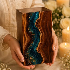 May include: A rectangular wooden box featuring a central vertical inlay of deep blue resin with swirling patterns and embedded gold leaf flakes. The wood displays a rich brown grain, and the resin creates a smooth, river-like effect.