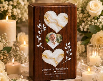 May include: A rectangular dark wood memorial urn featuring two white marble-like heart inlays with gold flecks and a central oval photo frame of a man. White vine illustrations flank the hearts, with the text Forever in Our Hearts, Always Loved, Never Forgotten, 1955-2024.