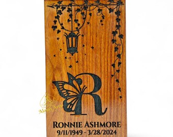 May include: Wooden urn with a unique design. The front features a large letter "R" with a butterfly wing, a lantern, and the name "RONNIE ASHMORE" with birth and death dates. The wood has a warm, natural grain.