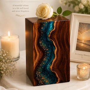 May include: A rectangular wooden cremation urn featuring a central vertical inlay of blue resin with gold leaf accents. The dark wood grain contrasts with the smooth, flowing blue resin river design. A single white rose rests on the top surface.