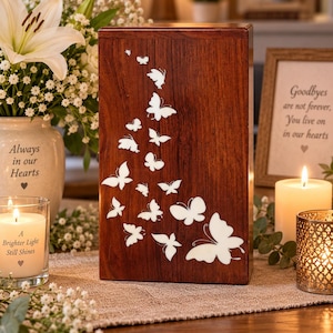 Op de afbeelding: Een rechthoekige houten urn met een donkerbruine afwerking, voorzien van een decoratieve inleg van meerdere witte vlinders van verschillende groottes, gerangschikt in een vloeiend patroon over de voorzijde van de rechtopstaande houten doos.