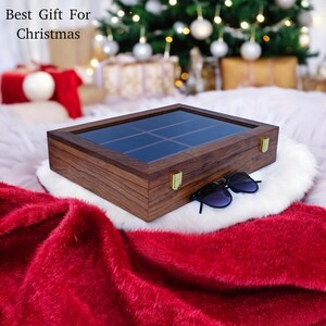 K&ouml;nnte beinhalten: Eine rechteckige Aufbewahrungsbox aus Holz mit Glasdeckel und goldfarbenen Metallverschl&uuml;ssen. Die Box hat eine dunkelbraune Holzmaserung und ist in neun F&auml;cher unterteilt. Eine schwarze Sonnenbrille liegt auf der wei&szlig;en Oberfl&auml;che.