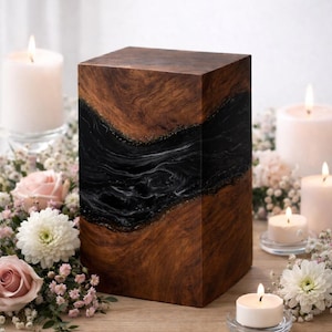 May include: A wooden urn with a dark, swirling black and white design. The wood has a rich, brown grain. The urn is rectangular and sits on a wooden surface, surrounded by candles and flowers.