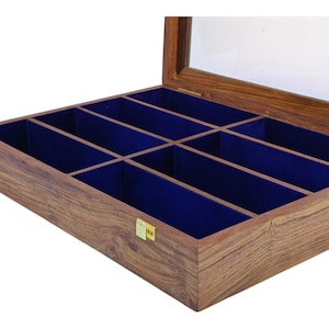 K&ouml;nnte beinhalten: Eine rechteckige Aufbewahrungsbox aus Holz mit einem aufklappbaren Glasdeckel und goldfarbenen Metallverschl&uuml;ssen. Der Innenraum ist in acht separate F&auml;cher unterteilt, die jeweils mit einem tiefblauen Stoff ausgekleidet sind.