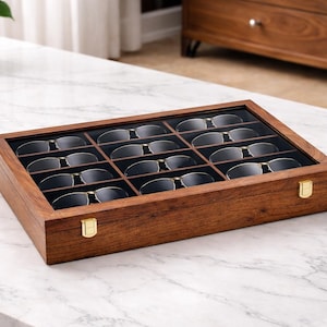 K&ouml;nnte beinhalten: Eine rechteckige Aufbewahrungsbox aus Holz mit einem klaren Glasdeckel, die zw&ouml;lf einzelne F&auml;cher zur Aufbewahrung von Brillen enth&auml;lt. Die Box besteht aus dunkelbraunem Holz und hat zwei goldfarbene Metallverschl&uuml;sse an der Vorderseite.