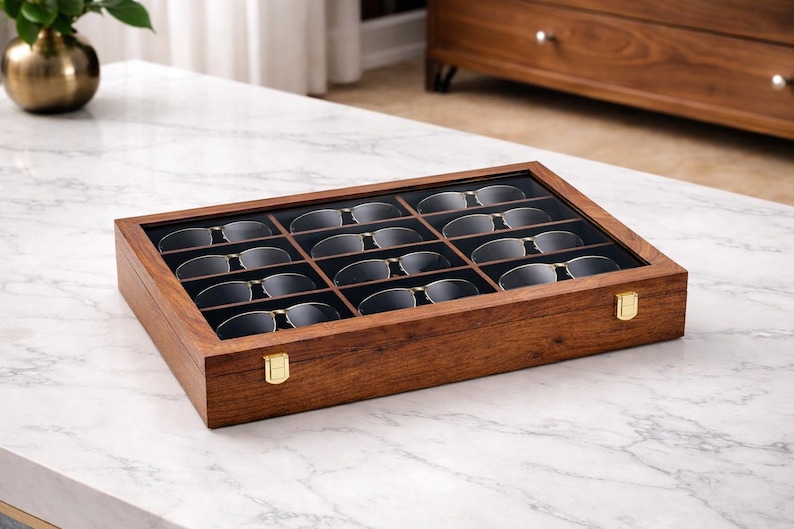 K&ouml;nnte beinhalten: Eine rechteckige Aufbewahrungsbox aus Holz mit einem klaren Glasdeckel, die zw&ouml;lf einzelne F&auml;cher zur Aufbewahrung von Brillen enth&auml;lt. Die Box besteht aus dunkelbraunem Holz und hat zwei goldfarbene Metallverschl&uuml;sse an der Vorderseite.