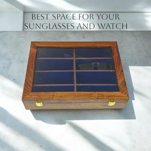 K&ouml;nnte beinhalten: Eine rechteckige Aufbewahrungsbox aus Holz mit Glasdeckel und zwei goldfarbenen Metallverschl&uuml;ssen. Das Innere ist mit dunkelblauem Samt ausgekleidet und in zehn F&auml;cher unterteilt, in denen eine schwarze Sonnenbrille liegt. Text: BEST SPACE FOR YOUR SUNGLASSES AND WATCH.