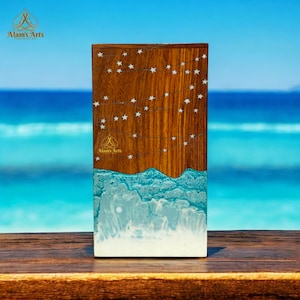 May include: A rectangular wooden art piece with a star design. The top portion is brown wood with white stars. The bottom section has a blue and white wave design. The background is a blurred ocean scene.