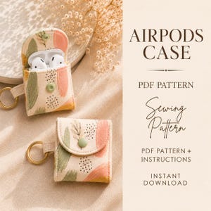 Op de afbeelding: Twee stoffen AirPods-hoesjes met een crèmekleurige achtergrond, abstracte penseelstreken in saliegroen, perzik en geel, geaccentueerd met zwarte stippen en bladmotieven. Elk hoesje heeft een goudkleurige sleutelhanger en een groene drukknoop.