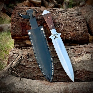May include: A hunting knife with a polished steel blade and a wooden handle with a silver bolster. The knife is next to a black leather sheath with white stitching. The knife and sheath are resting on a weathered log.