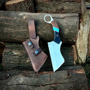 May include: A curved blade knife with a wooden handle, turquoise accents, and a black base. The knife is paired with a brown leather sheath with two snap closures. The knife and sheath are set against a backdrop of stacked logs.