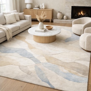 Peut inclure: Un grand tapis avec un motif abstrait aux tons crème, beige et bleu clair. Le tapis est dans un salon, avec un canapé, des chaises et une table basse. Le motif du tapis évoque l'eau qui coule.