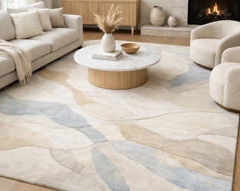 May include: A large area rug with a flowing, abstract design in shades of cream, beige, and light blue. The rug is placed in a living room setting, with a sofa, chairs, and a coffee table visible.