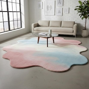 May include: A large, irregularly shaped rug with a soft, plush texture. The rug features a watercolor design with blended hues of pink, blue, and white. It is placed on a concrete floor in a living room setting.