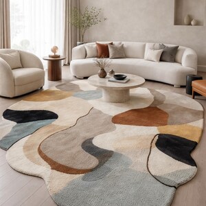 Op de afbeelding: Een groot, abstract vloerkleed met een unieke, organische vorm. Het vloerkleed heeft een palet van neutrale kleuren, waaronder beige, bruin, grijs en zwart, wat een moderne en stijlvolle uitstraling creëert. Het vloerkleed bevindt zich in een woonkamer met een witte bank en stoelen.