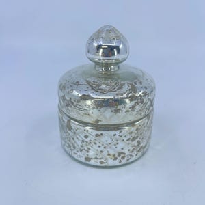 May include: A silver-colored, decorative jar with a lid. The jar has a round body with a diamond pattern and a distressed finish. The lid is a rounded shape with a small knob on top. The jar is likely made of glass or a similar material.