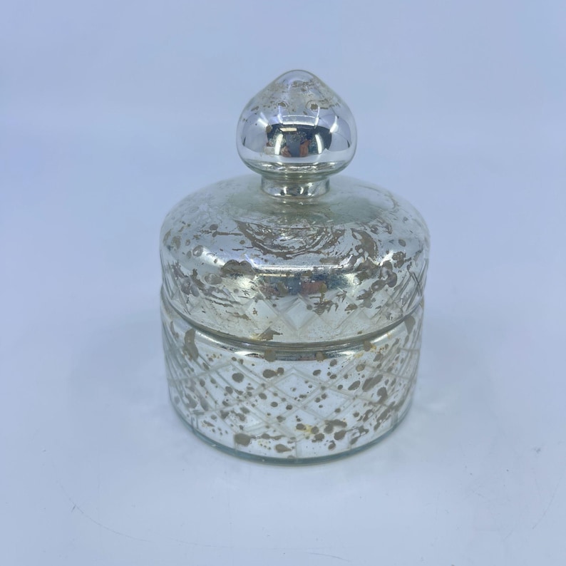 May include: A silver-colored, decorative jar with a lid. The jar has a round body with a diamond pattern and a distressed finish. The lid is a rounded shape with a small knob on top. The jar is likely made of glass or a similar material.