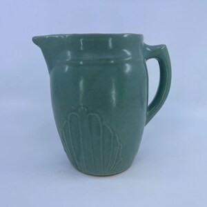 May include: A sage green ceramic pitcher featuring a smooth, matte finish and a molded shell-like relief pattern on the lower body. The vessel includes a curved handle on the right side and a prominent pouring spout at the top rim of the container.