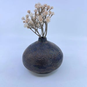 May include: A dark blue, spherical ceramic vase with a textured, mottled finish and an etched floral pattern around the center. The vase contains a bouquet of small, light beige artificial flowers with thin, branching stems extending upward.