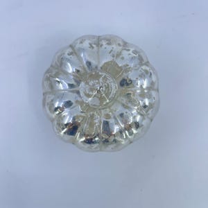 May include: A silver-coloured, decorative pumpkin ornament with a textured, distressed finish. The pumpkin has a ribbed design and a circular indentation at the top. The ornament is likely for autumn or Thanksgiving decor.