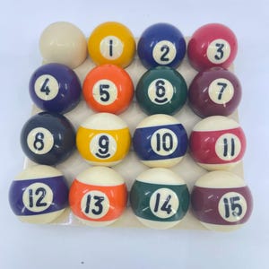 May include: A complete set of fifteen numbered billiard balls and one white cue ball arranged in a grid. The set includes solid colored balls numbered one through seven and striped balls numbered nine through fifteen, all featuring black numerals.