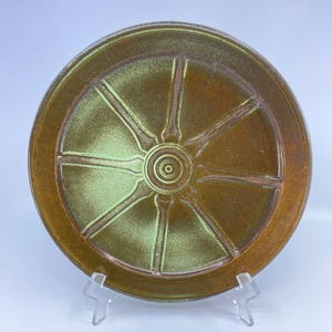 May include: A round, ceramic plate with a wheel-like design. The plate features a central circular indentation surrounded by radiating lines, creating a segmented pattern. The color is a blend of brown and green, with a glossy finish.