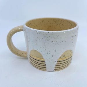 May include: A white ceramic mug with a speckled exterior and a tan interior. The mug features a tan handle and a decorative design of tan arches with gold horizontal lines. The mug is approximately 4 inches tall.
