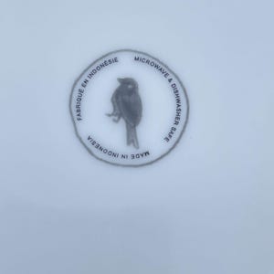 May include: A white ceramic plate with a circular design featuring a bird illustration. The text around the bird reads "Fabriqu&eacute; en Indon&eacute;sie", "Microwave & Dishwasher Safe", and "Made in Indonesia".