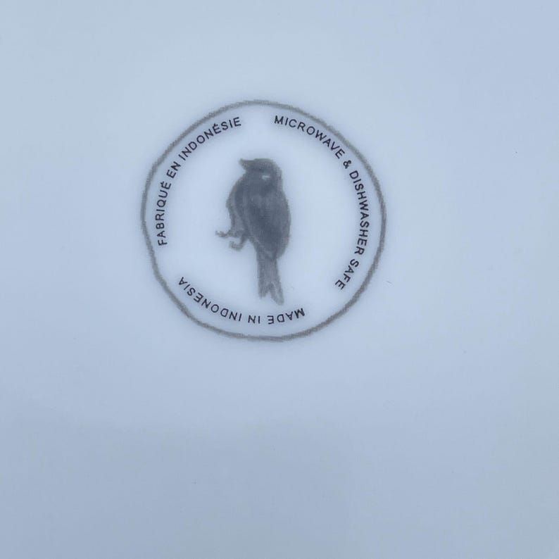 May include: A white ceramic plate with a circular design featuring a bird illustration. The text around the bird reads "Fabriqu&eacute; en Indon&eacute;sie", "Microwave & Dishwasher Safe", and "Made in Indonesia".