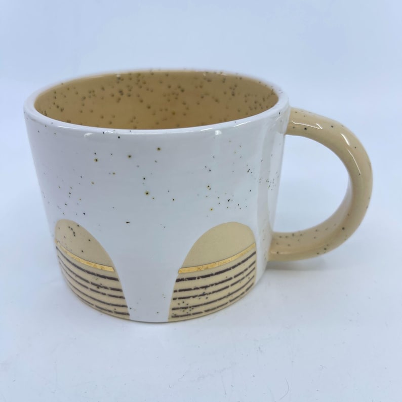 May include: White ceramic mug with a speckled tan interior and handle. The exterior features a white base with tan and gold geometric designs. The mug has a rounded shape and a comfortable handle.